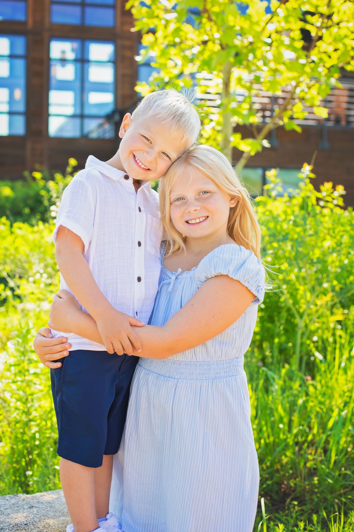 Smiling blonde girl embraces her younger brother outdoors in front of a building with green foliage.