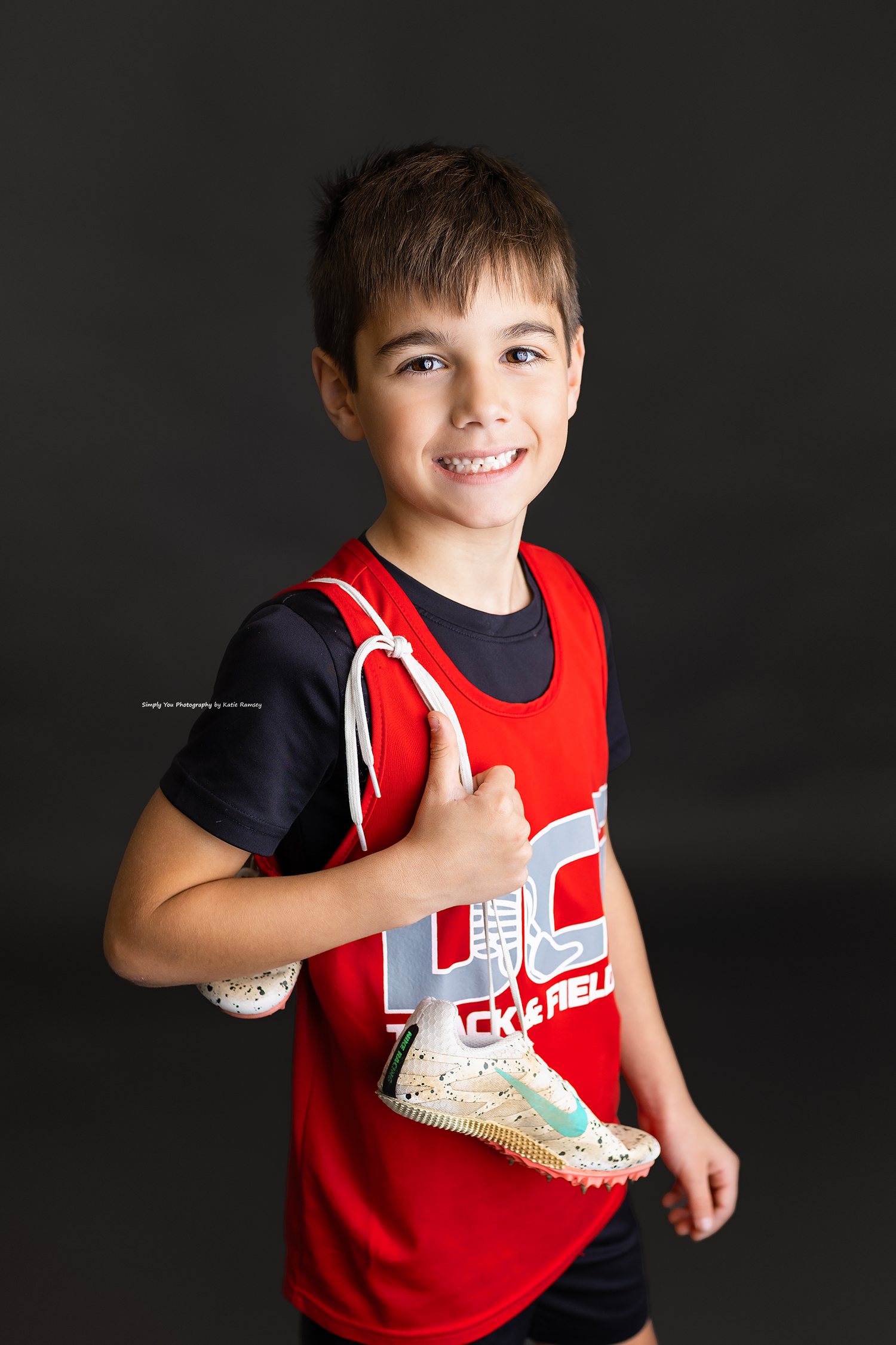 Smiling boy in red track uniform gives thumbs up while holding running cleats.