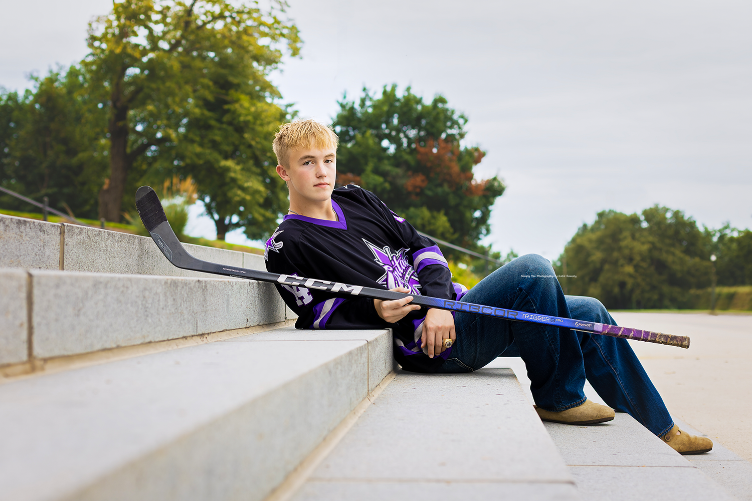 Young hockey player in team jersey sitting on steps outdoors, holding a purple and black hockey stick.