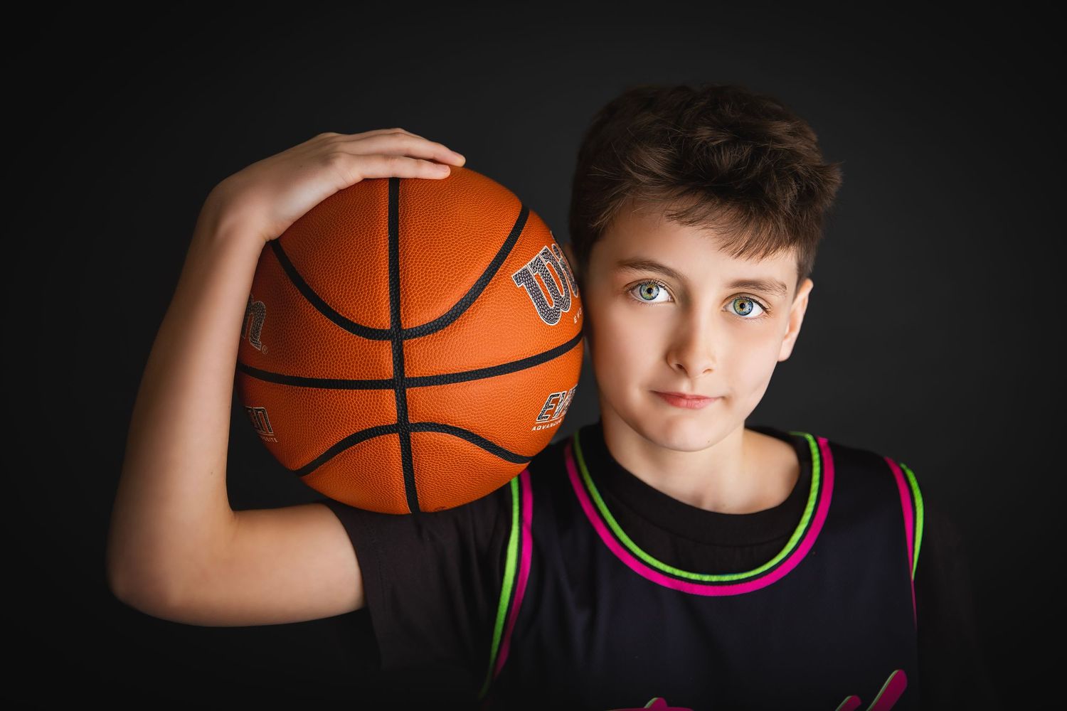 Young boy in black basketball jersey holding a Wilson basketball beside his head.