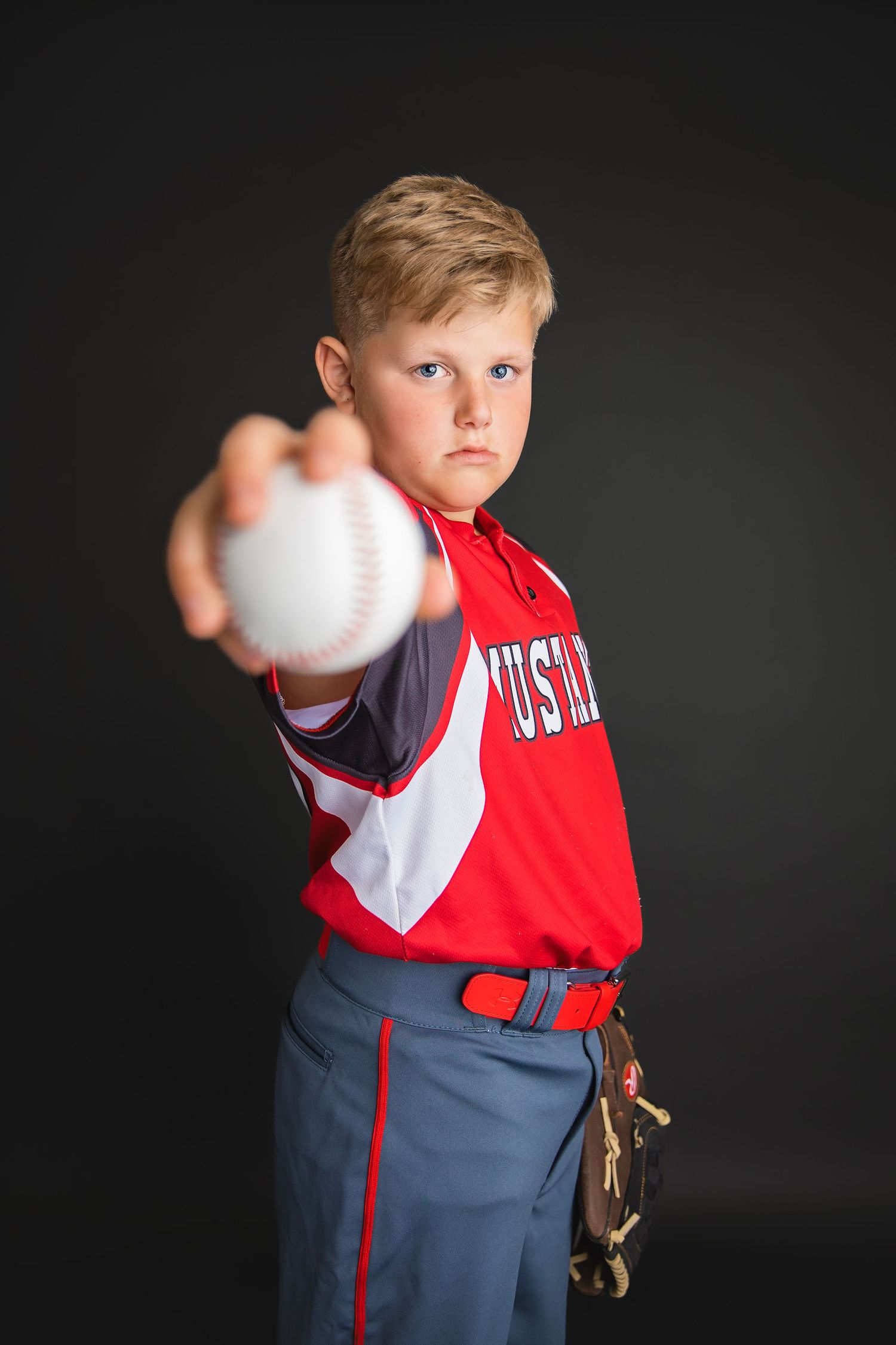 Determined young baseball player in red Mustangs uniform extends arm forward holding baseball toward camera.