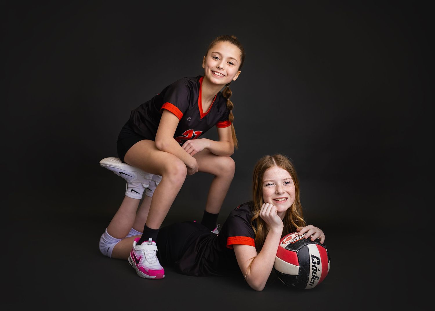 Two young girls in black and red volleyball uniforms pose together in a studio, one sitting elevated and one lying down with a ball.
