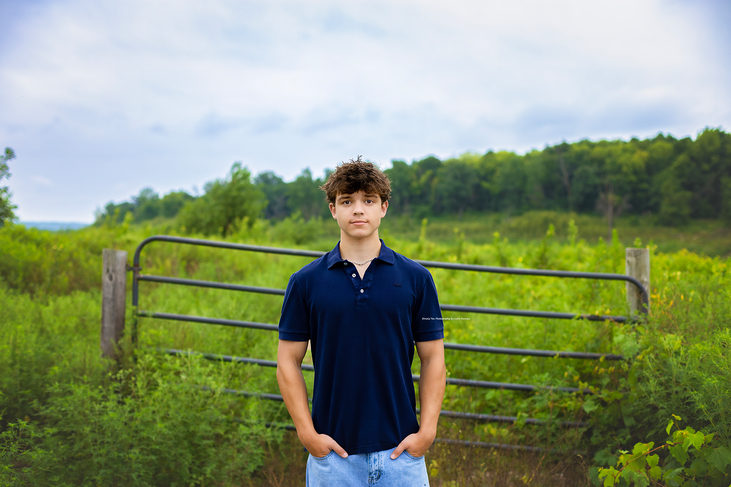 A young man in a navy polo shirt stands confidently in front of a metal gate surrounded by lush green fields.
