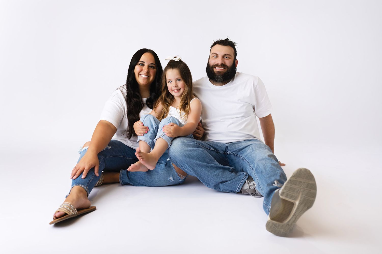 Happy family of four sitting together on white background, parents in white tees and jeans with two young children.