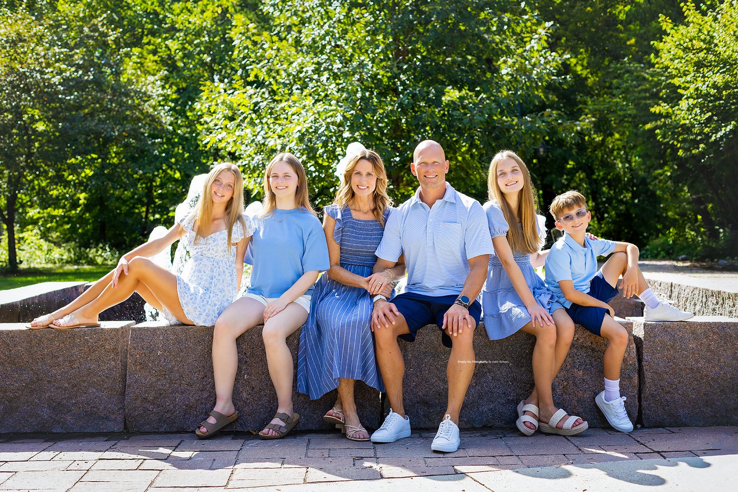A happy family of six poses outdoors on a stone bench surrounded by lush green trees on a sunny day.
