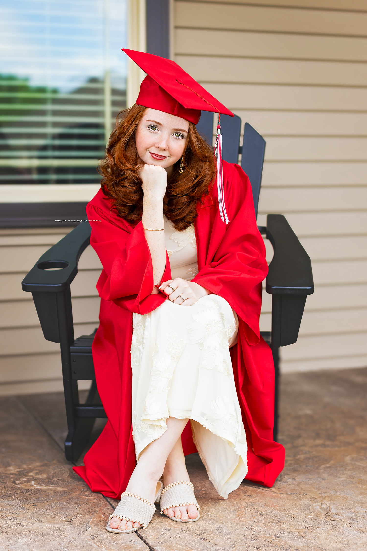 Redhead graduate sitting in black chair wearing red cap and gown over white dress with heels.