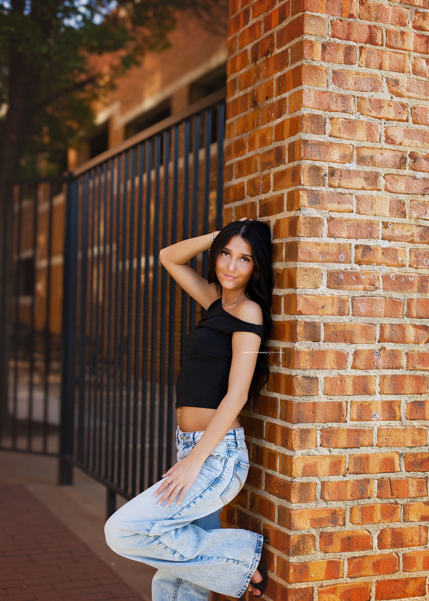 Young woman in black crop top and jeans leaning against a brick wall by a metal fence outdoors.