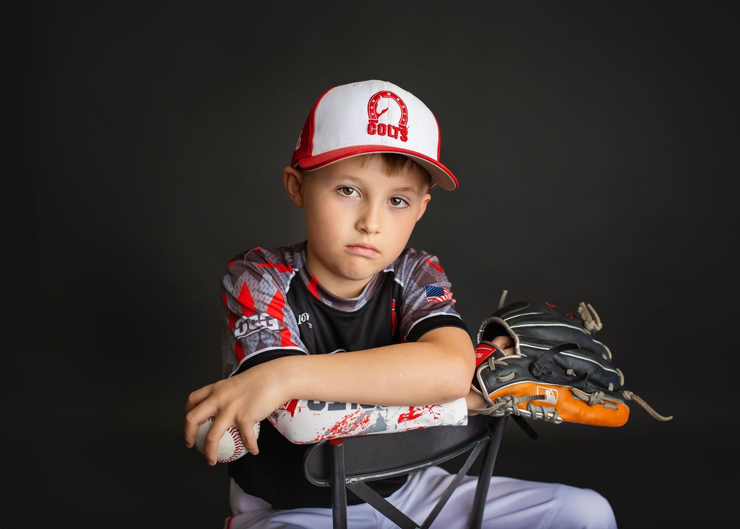 Young baseball player in red and white cap leans on bat with glove in studio portrait.