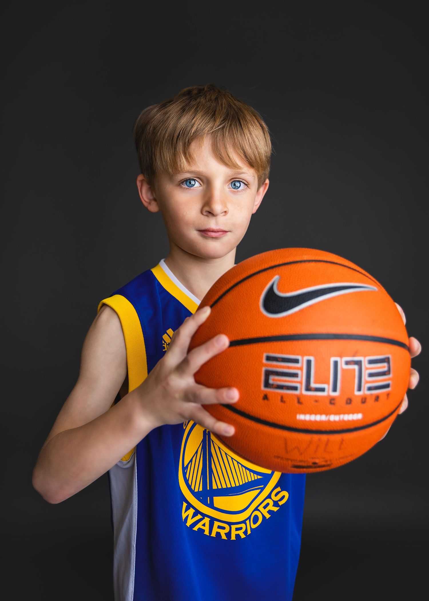 Blonde boy in Golden State Warriors jersey holds Nike Elite basketball forward.