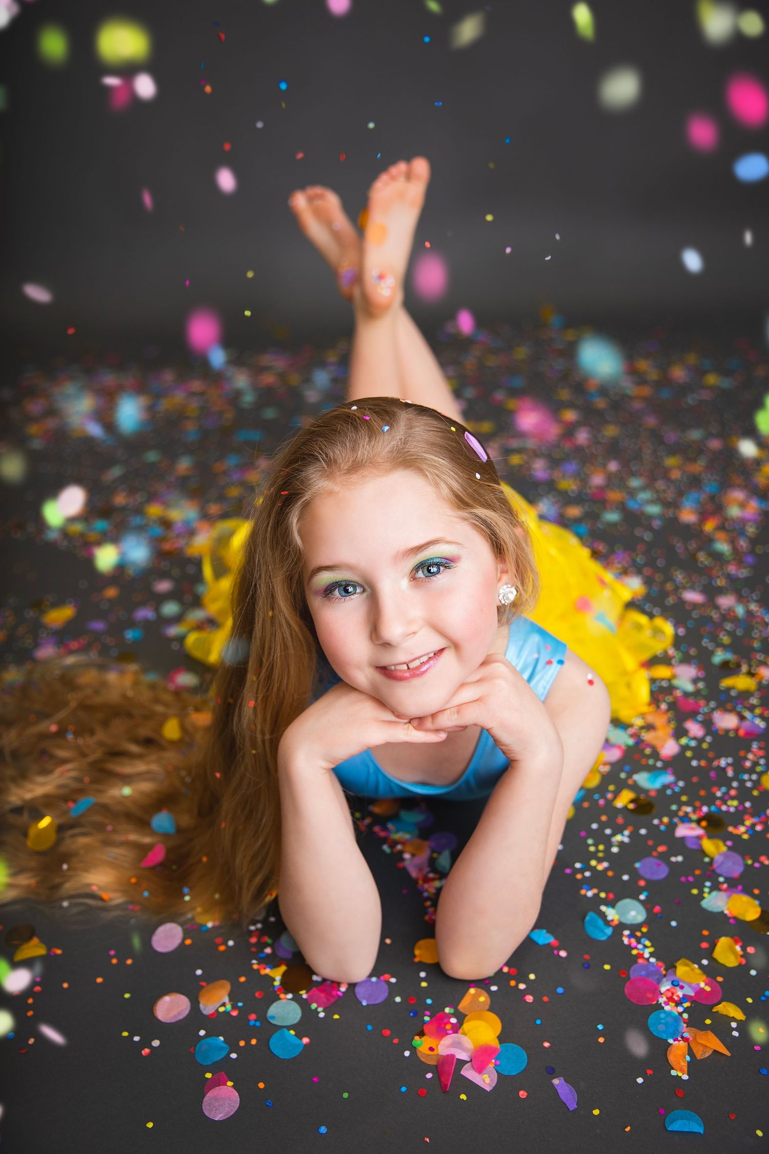 A smiling young girl in a blue top and yellow tutu lies on the floor surrounded by colorful confetti pieces.