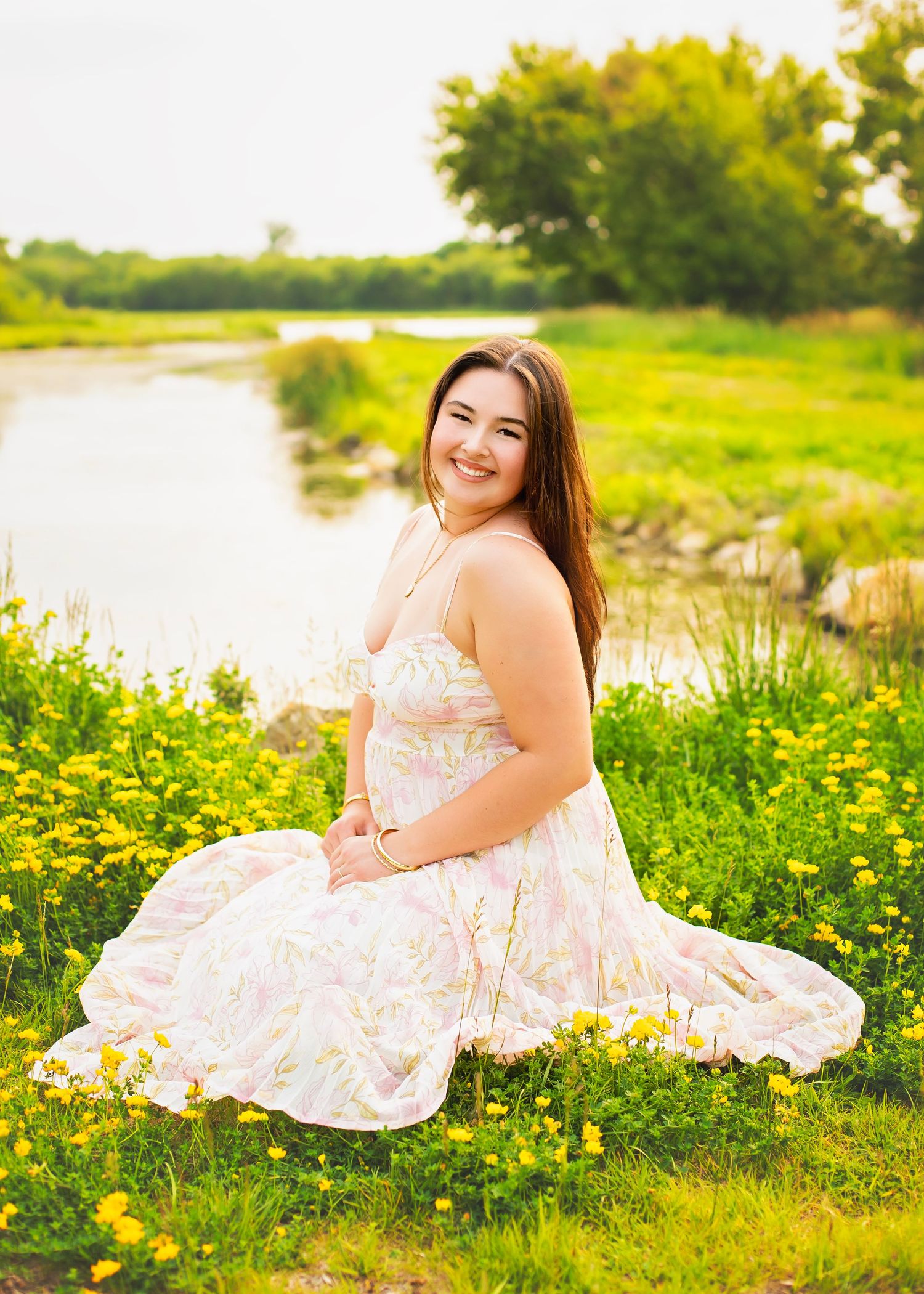 Young woman in floral dress sitting among yellow wildflowers near a pond.