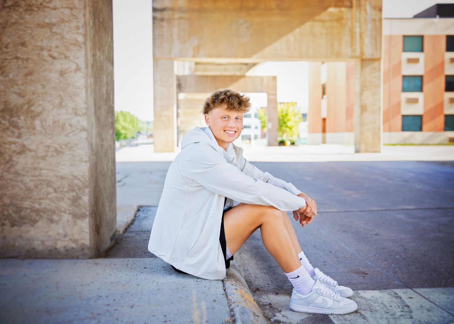 Smiling teen boy in gray outfit sitting on curb leaning against concrete pillar in urban setting.