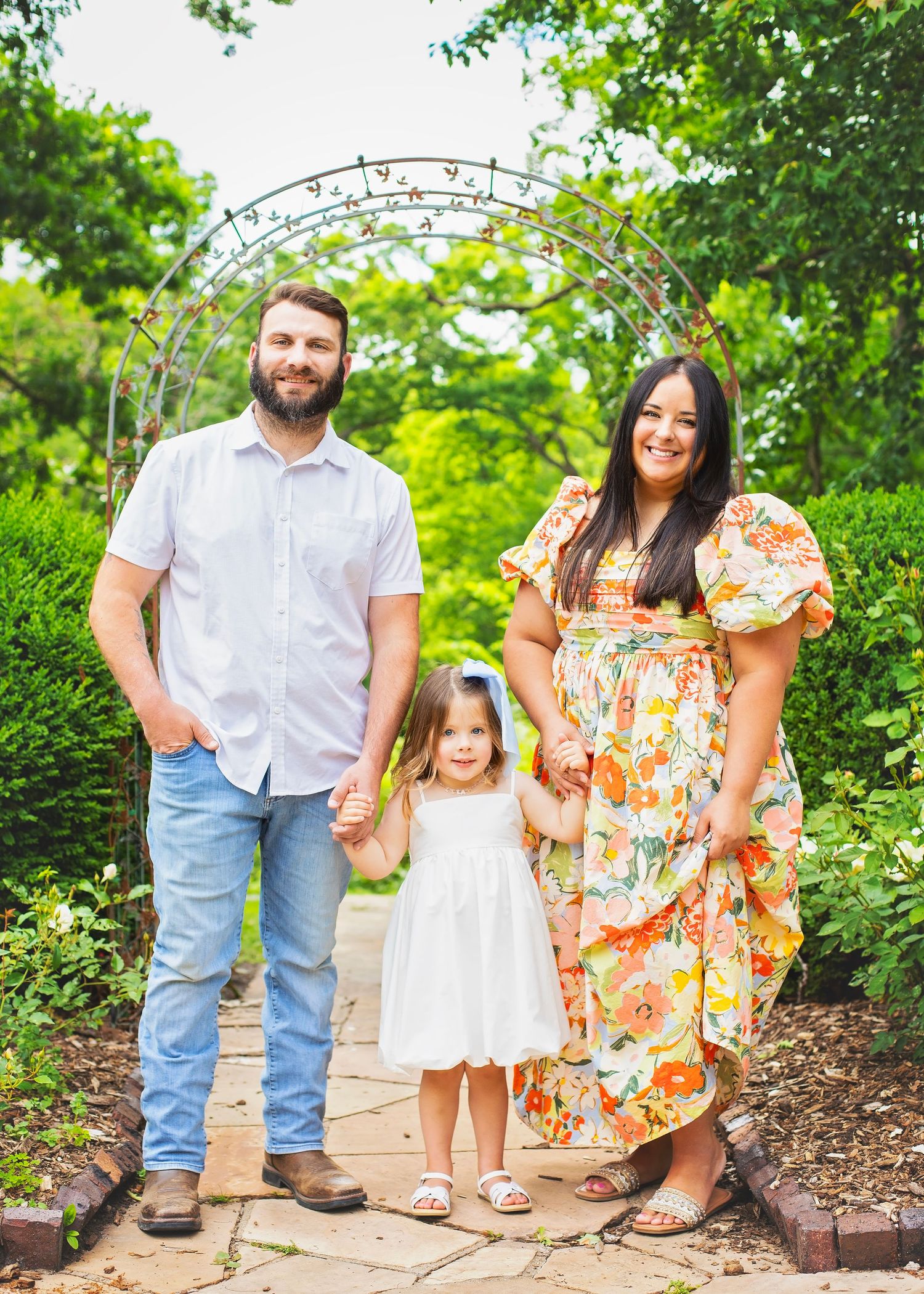 Happy family of three posing under a garden arch, parents holding hands with young daughter in white dress.