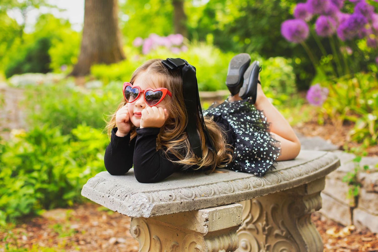 Cute little girl in black outfit and red heart sunglasses posing on stone bench in garden.