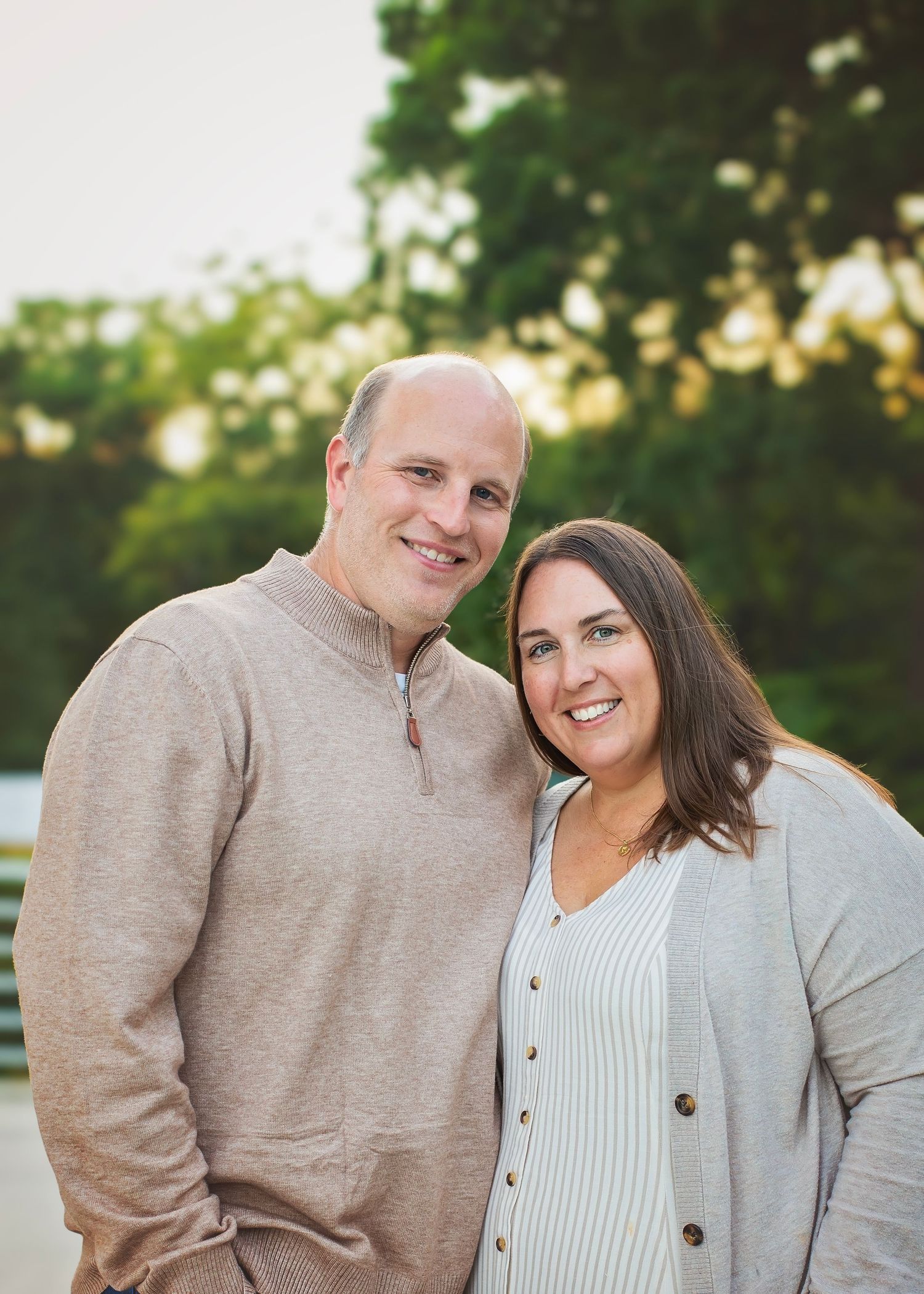 Smiling couple posing closely together outdoors with green trees and warm golden light in the background.
