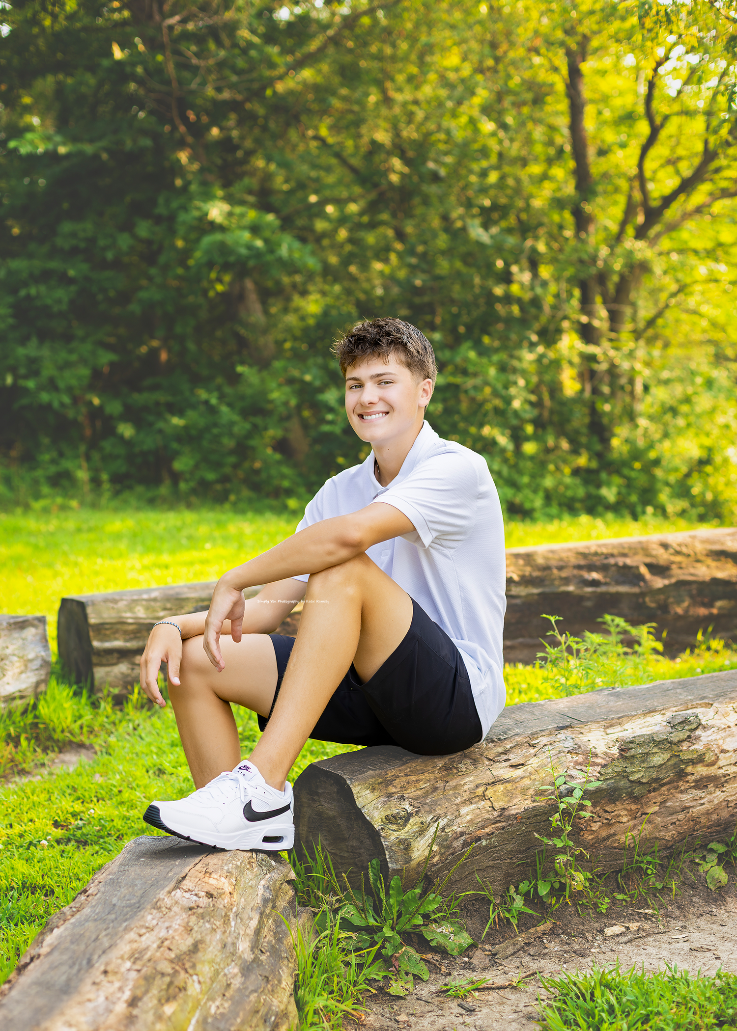 A smiling teenage boy sits on a log in a sunny park, wearing a white shirt, black shorts, and Nike sneakers.