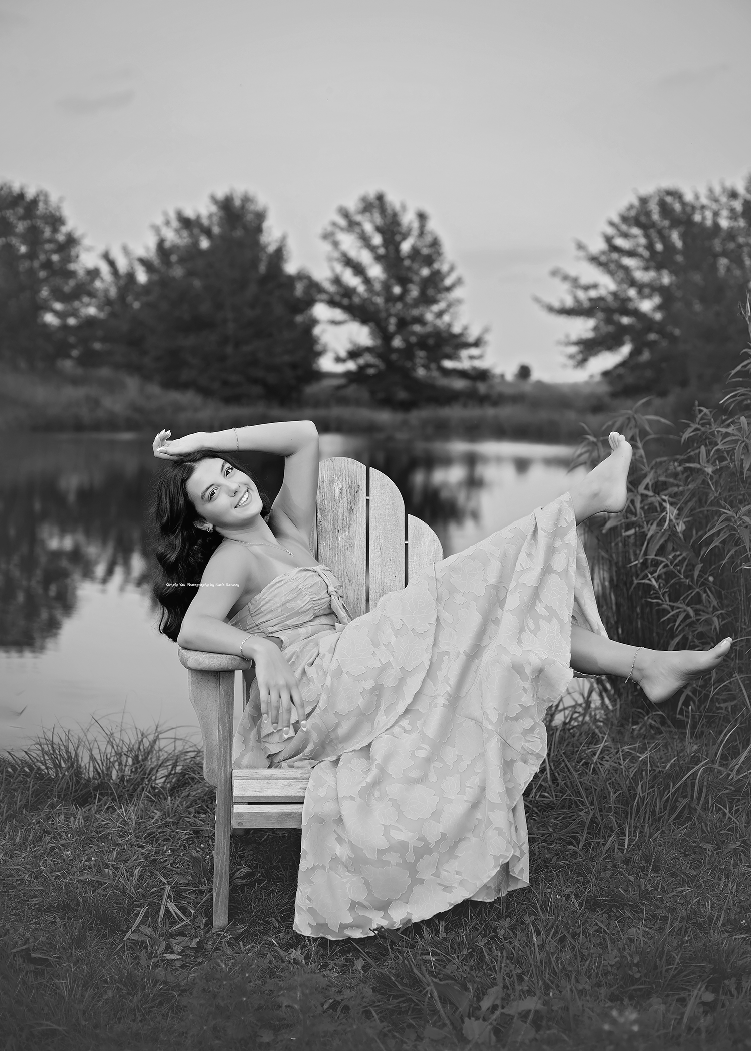 Black and white photo of a smiling woman in a flowy dress relaxing on an Adirondack chair by a serene pond.