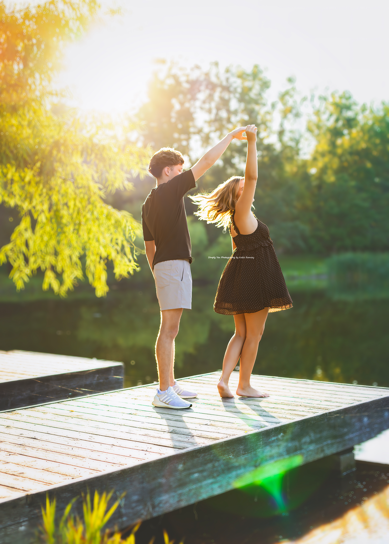 Couple dancing on a wooden dock surrounded by golden sunlight and lush green trees.