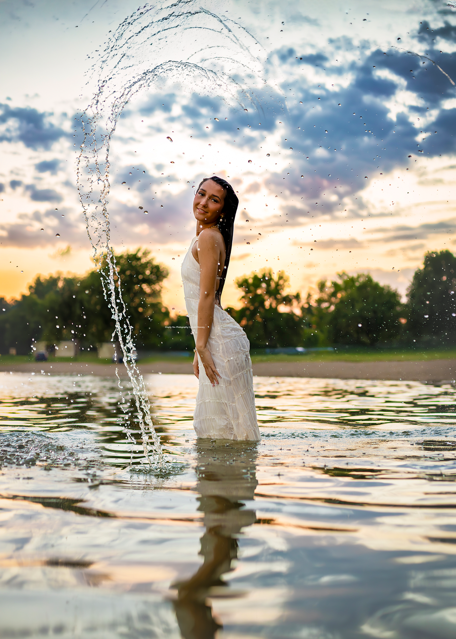 Woman in white dress standing in water with dramatic splash arc at sunset.