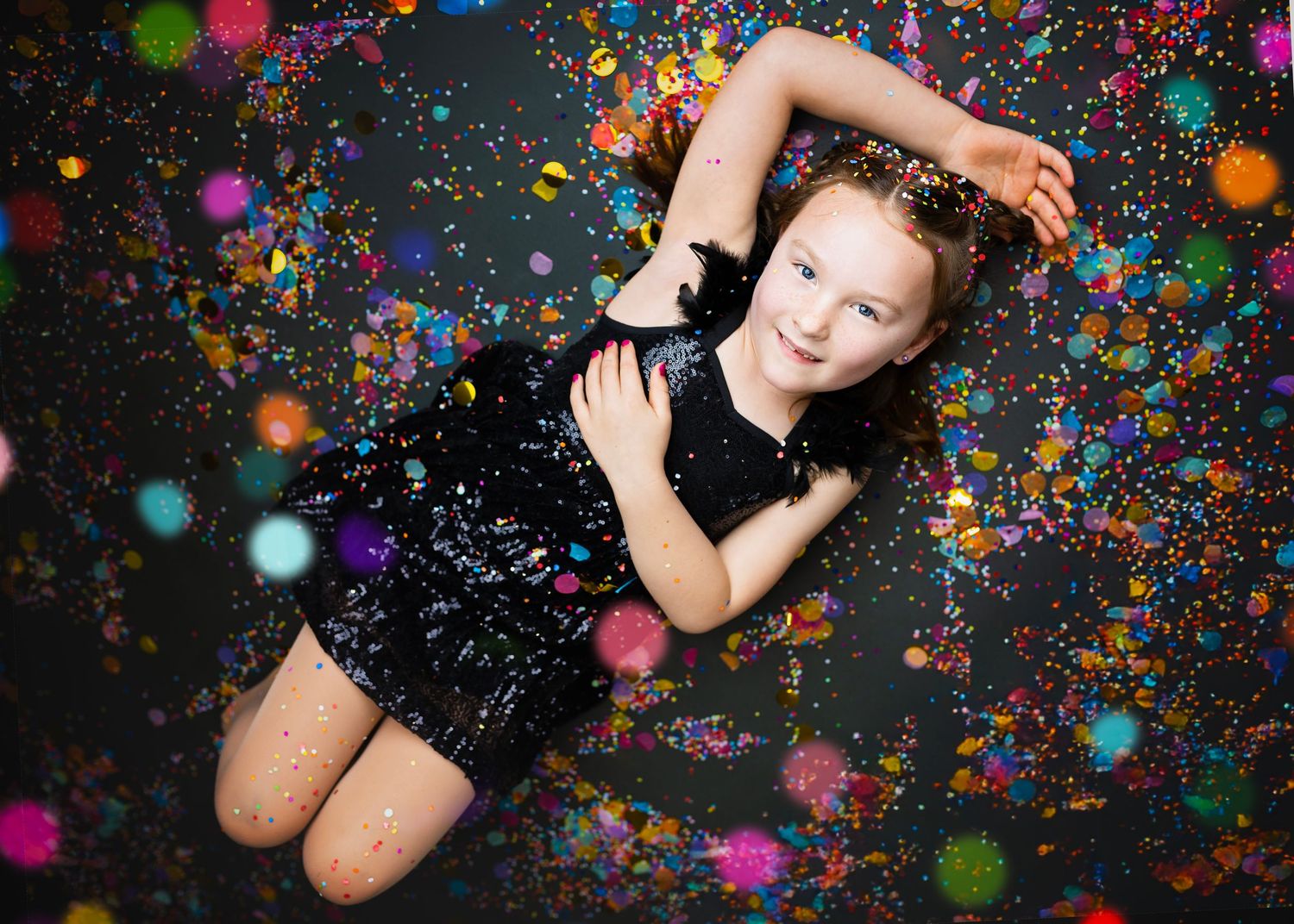 Young girl in black sequin dress posing on floor with vibrant confetti around her.