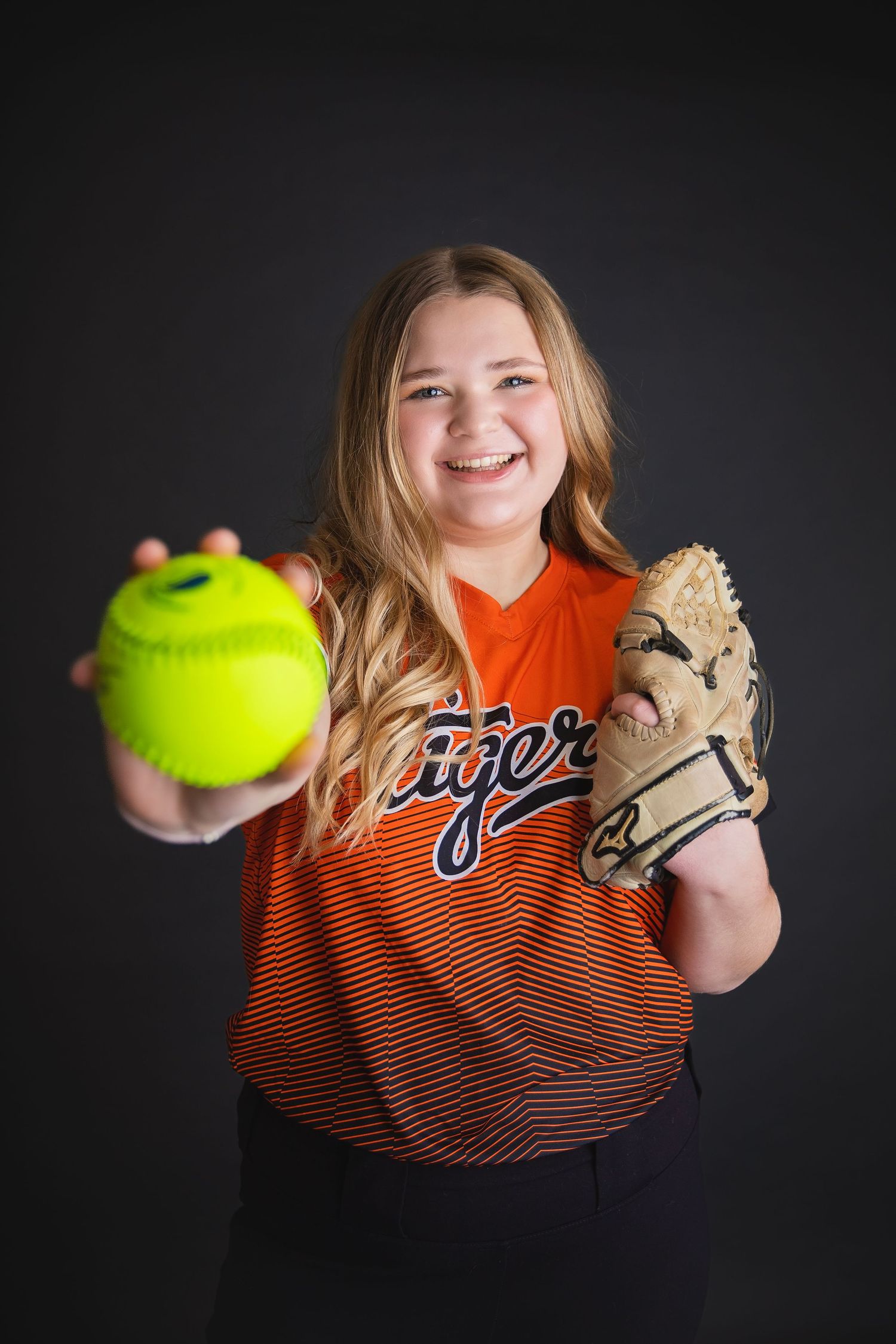 Young softball player in orange Tigers jersey holds yellow softball and glove toward camera.
