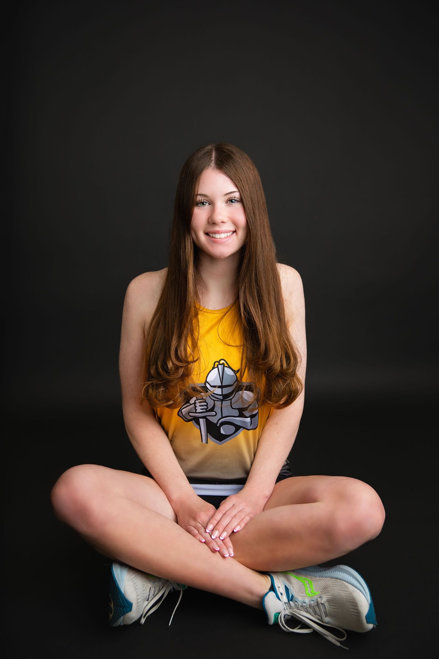 Smiling teenage girl in gold athletic tank top sits cross-legged on floor against dark background.