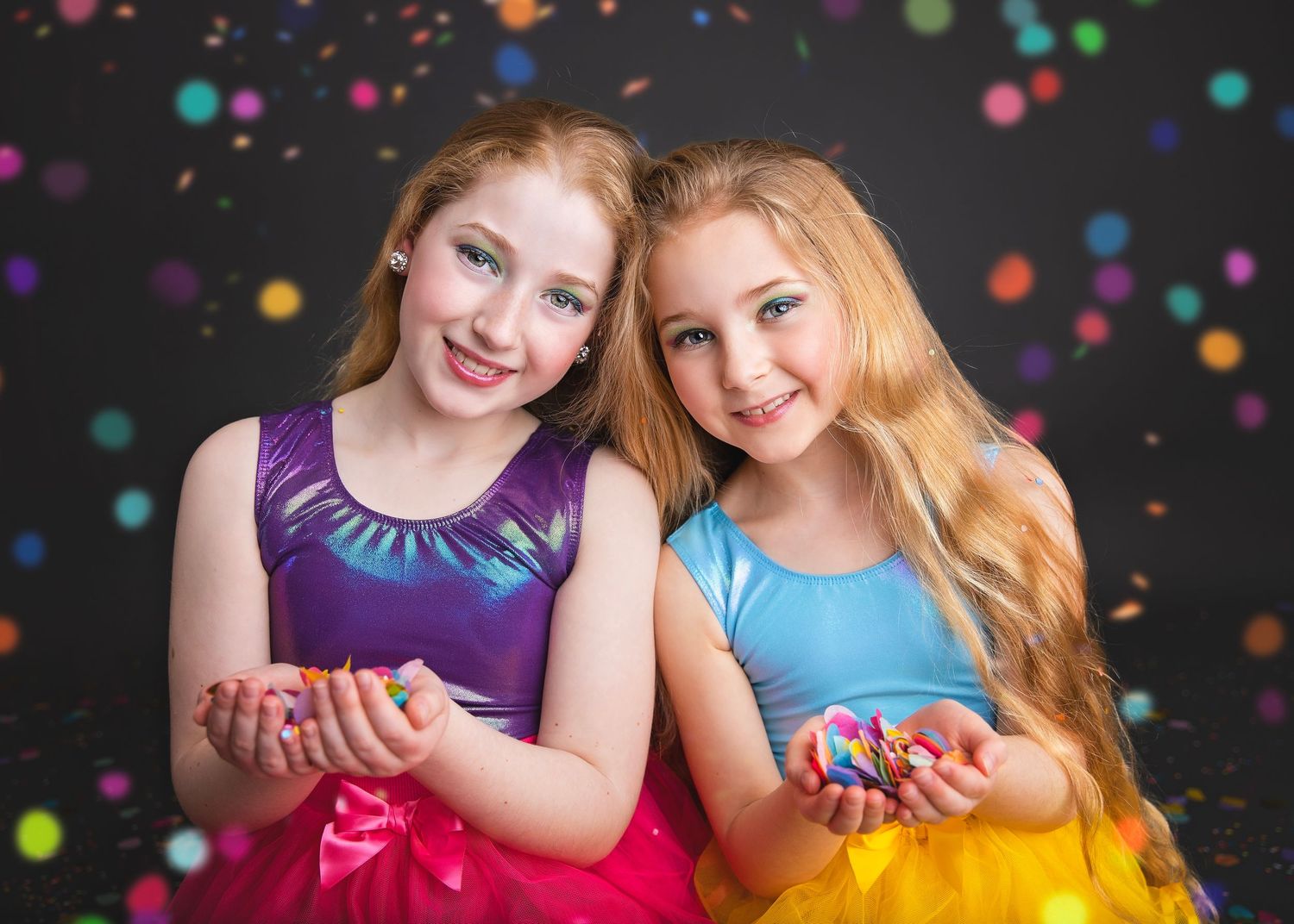 Two smiling girls in bright purple and yellow outfits holding colorful confetti with bokeh background.