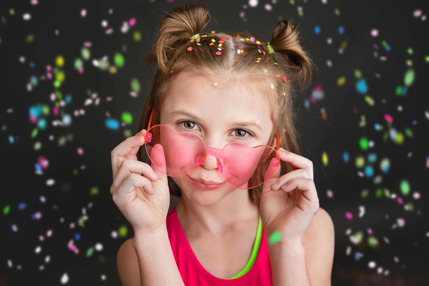 Playful girl in pink top adjusting red glasses with colorful confetti on dark background.