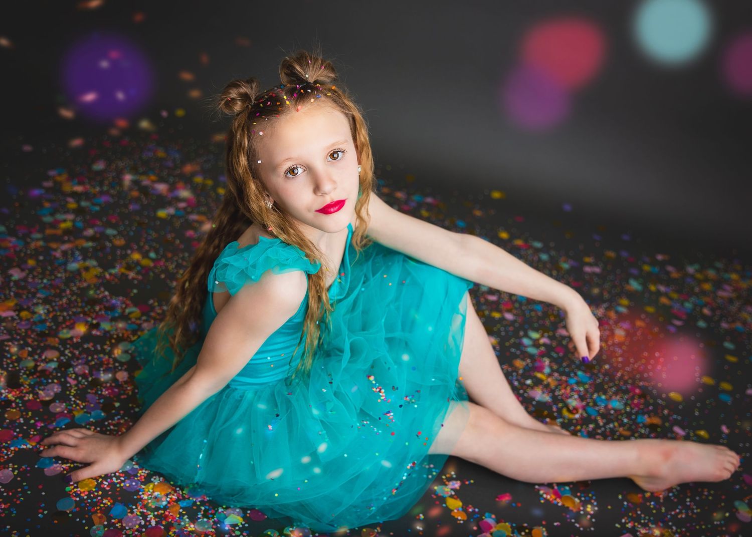 Girl in teal tulle dress sitting gracefully on confetti-covered studio floor.