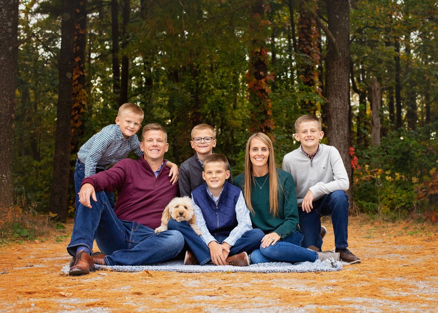 Family of six with a dog posing on a blanket in a pine forest during fall.