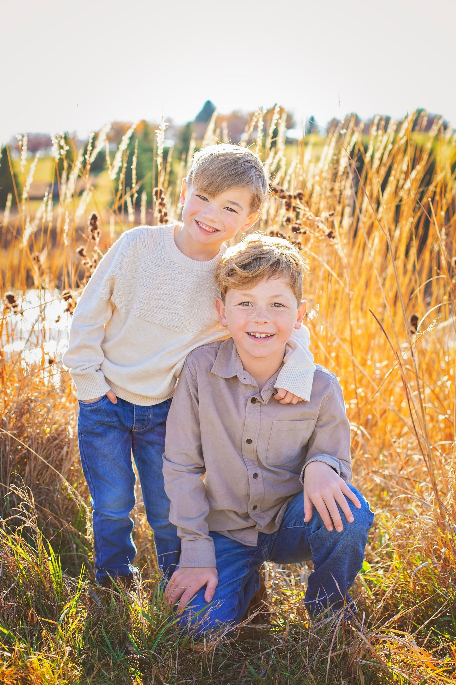 Two smiling blonde boys pose together in a golden autumn field, one crouching while the other stands behind.