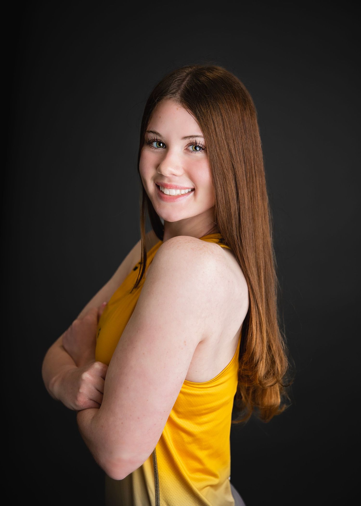 Smiling teenage girl in gold tank top with arms crossed posing sideways against dark background.