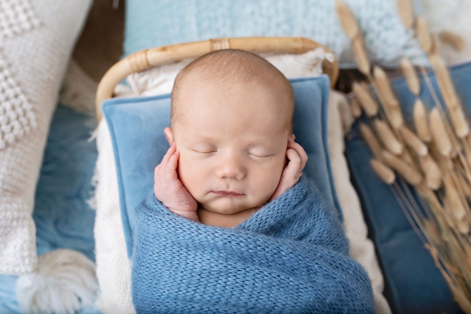 Sleeping newborn baby swaddled in blue wrap with hands framing face in wicker bassinet.