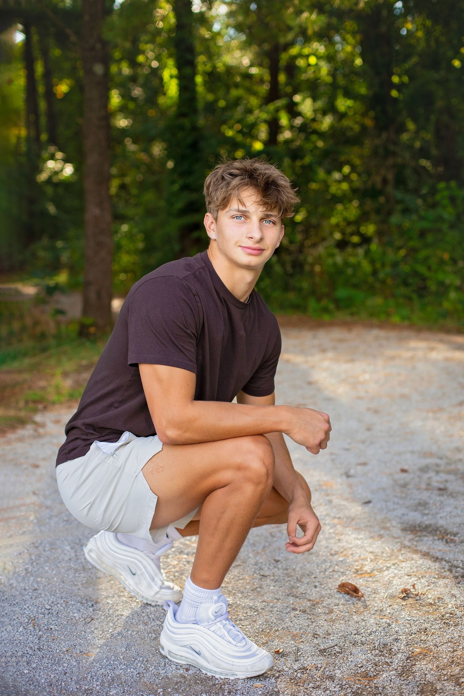 Young man in dark t-shirt crouches on a gravel path in a sunlit forest, wearing white sneakers.