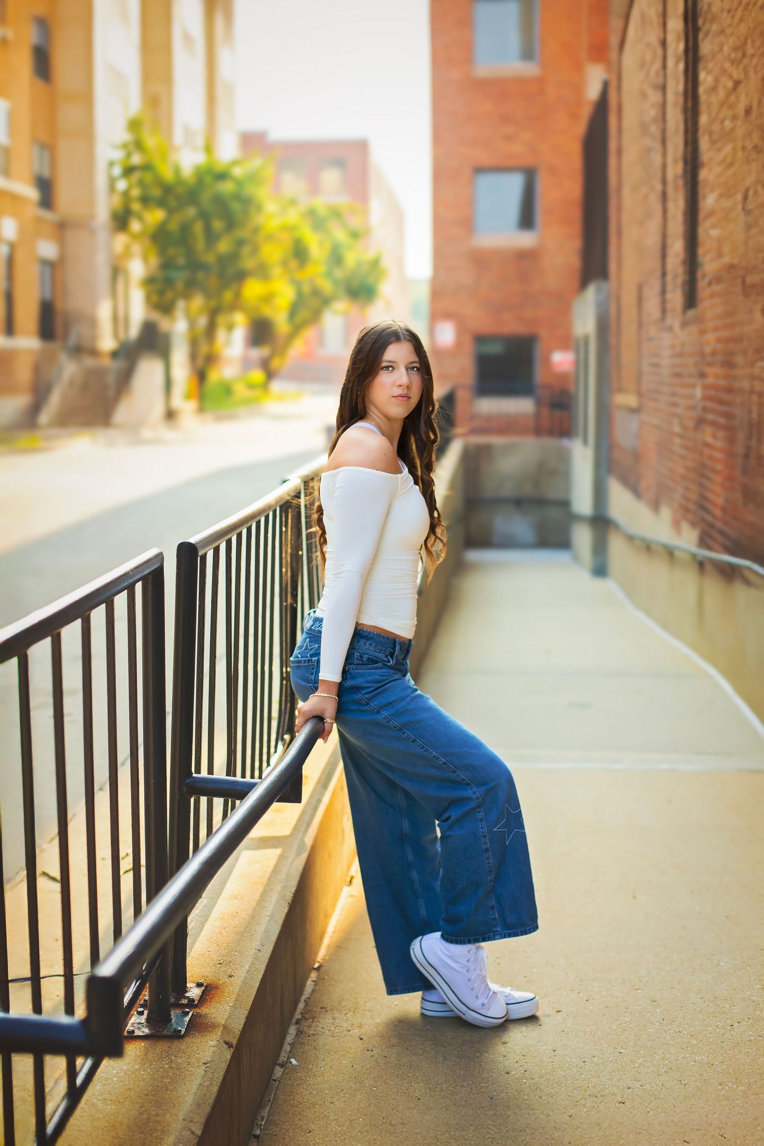 Young woman in white crop top and wide-leg jeans leans on railing in urban alleyway setting.