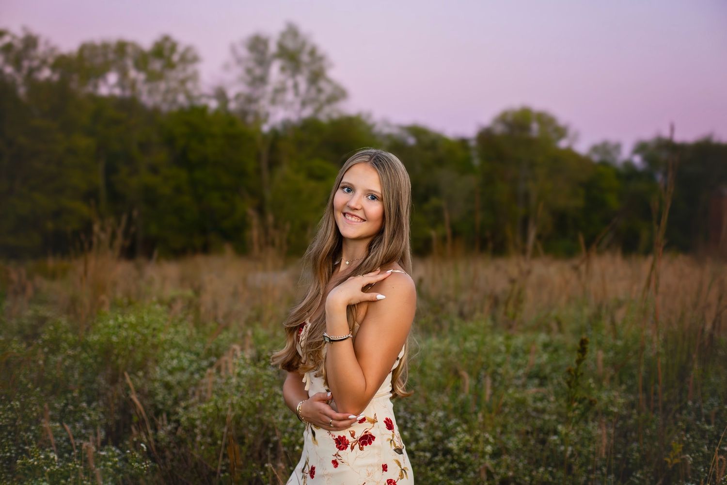Smiling brunette teen in floral dress poses in meadow under pink twilight sky at dusk.