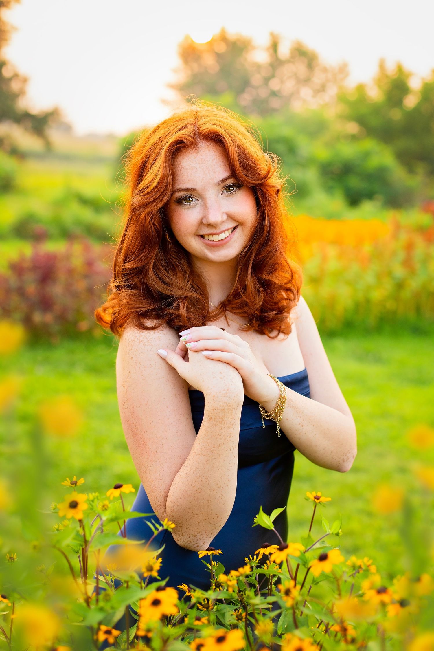 Smiling redhead teen with freckles in navy dress kneeling among yellow wildflowers.