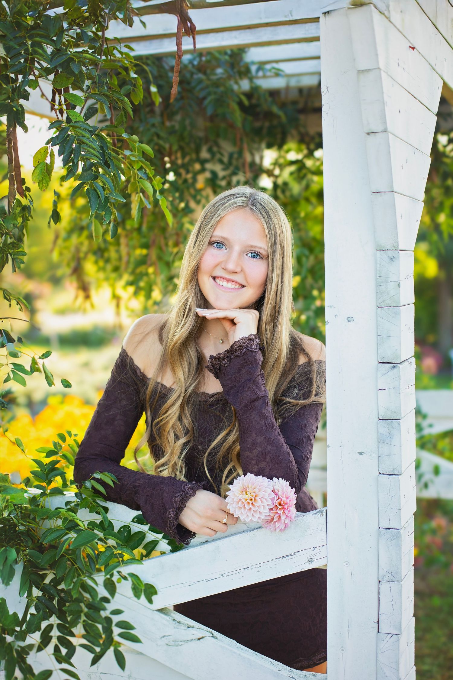 Smiling blonde teen in purple dress holds pink dahlia while leaning on white garden arbor railing.