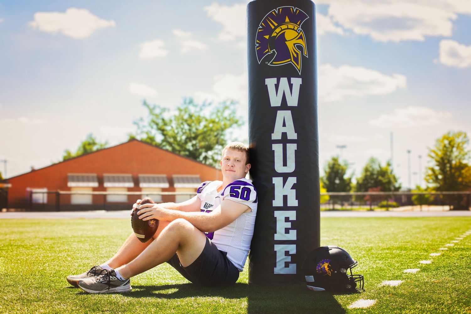 High school football player leaning against Waukee field goal post holding helmet on sunny day.