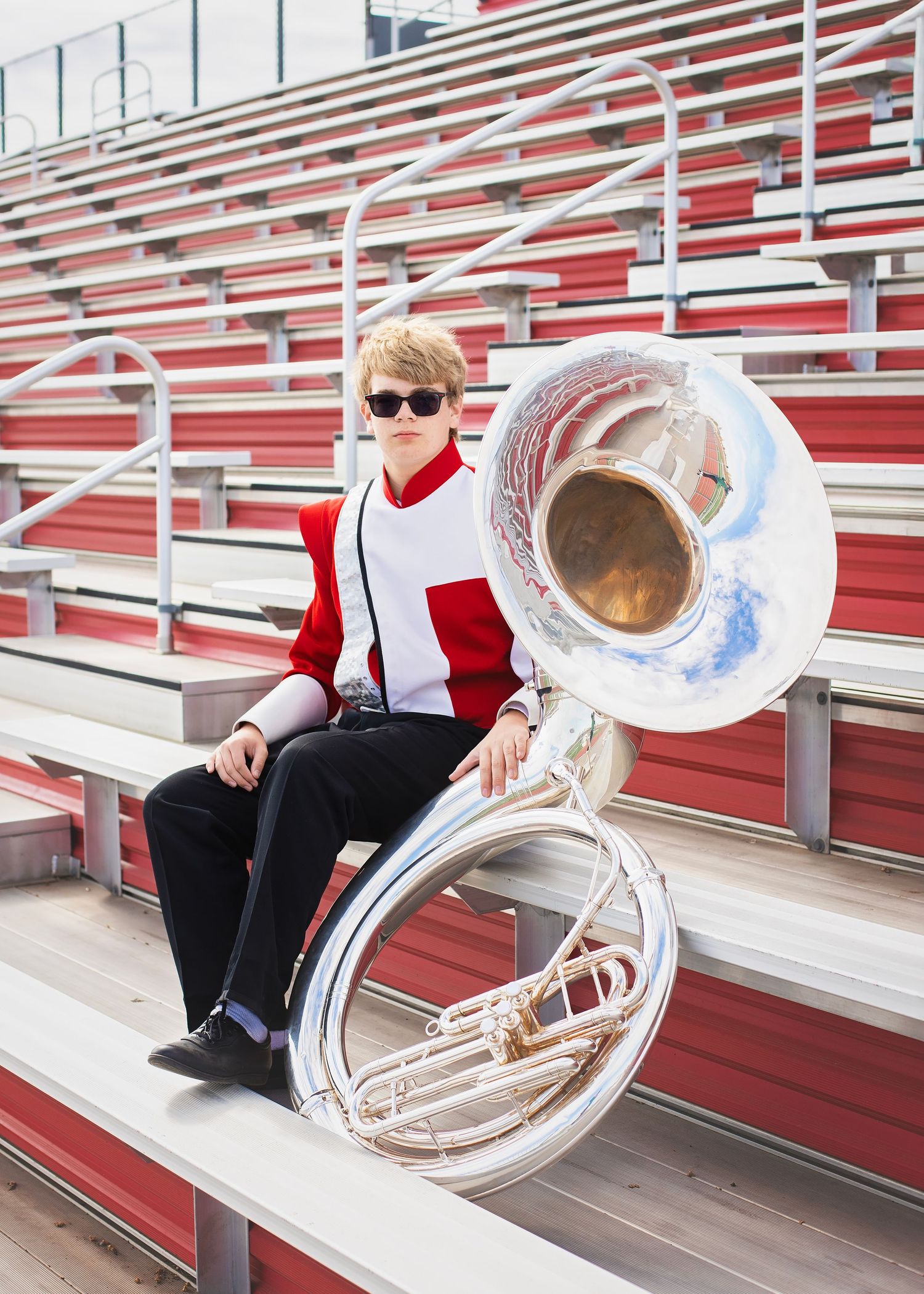 Teen in marching band uniform sitting in bleachers with a sousaphone at a football stadium.