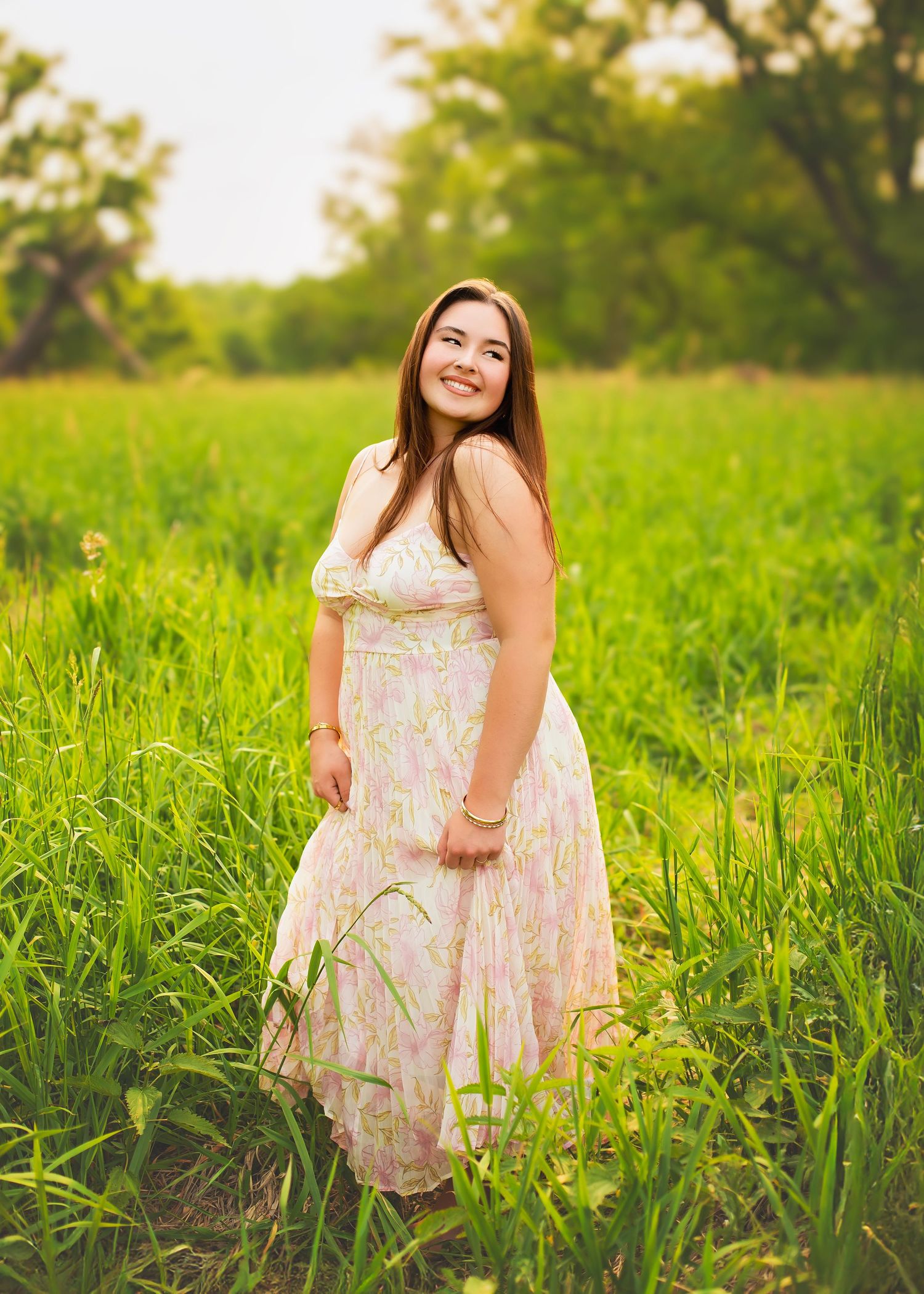 Young woman in pink floral sundress smiling in sunlit green meadow.