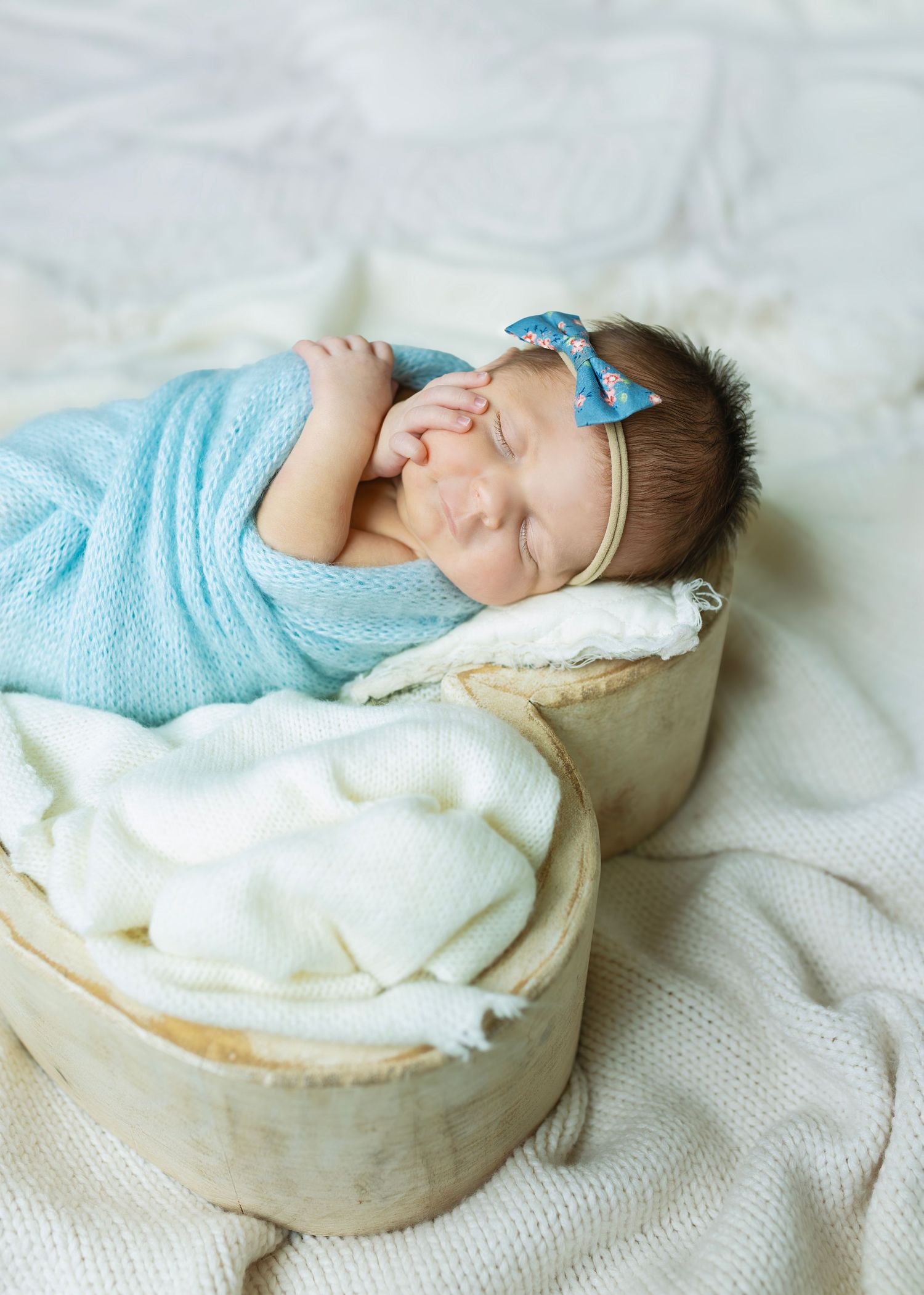 Sleeping newborn baby girl wrapped in blue knit swaddle with bow headband in heart-shaped bowl.