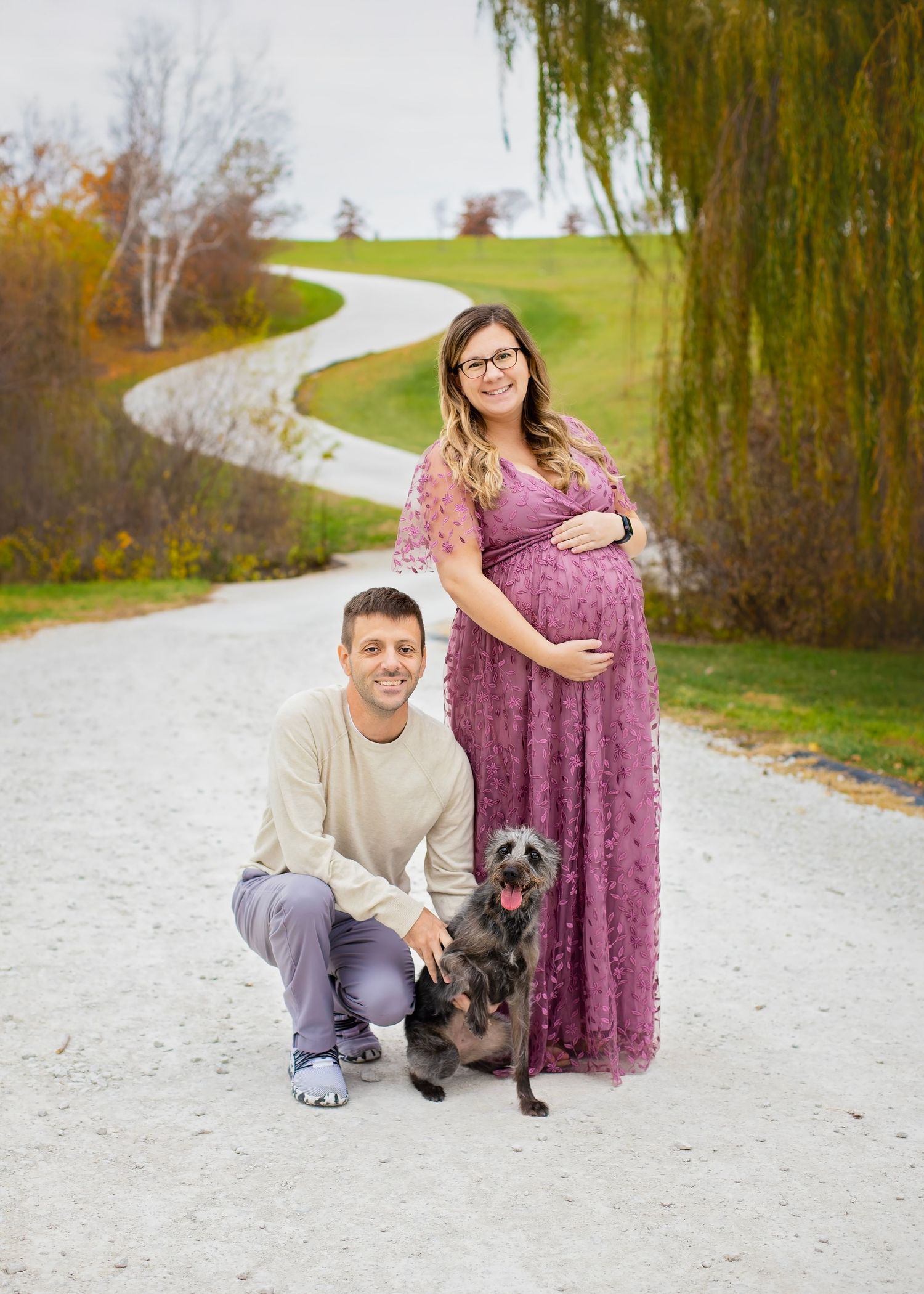Pregnant woman in purple lace gown stands with kneeling partner and small dog on winding path.
