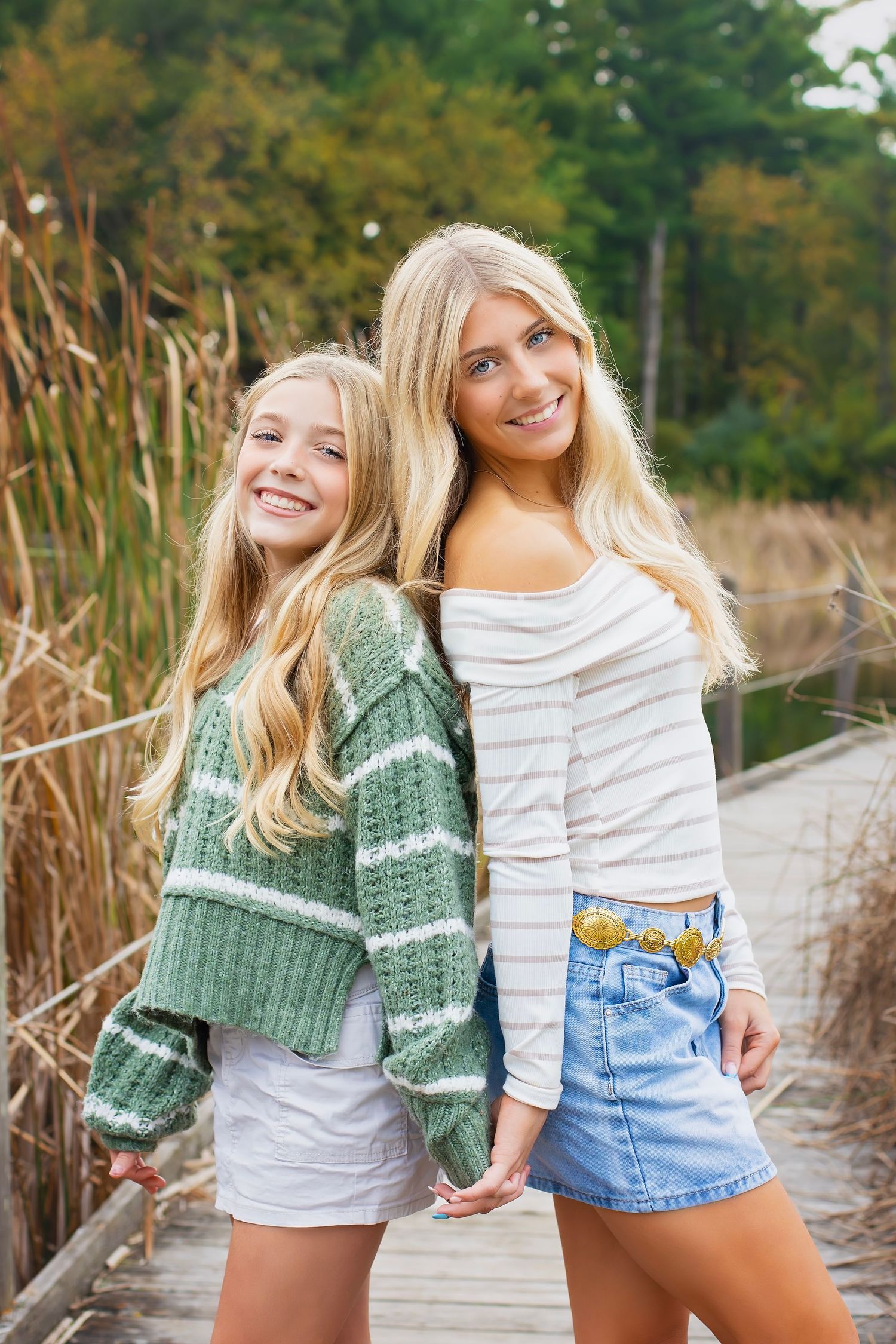 Two blonde sisters smile and pose back-to-back on a wooden boardwalk surrounded by tall autumn grasses.