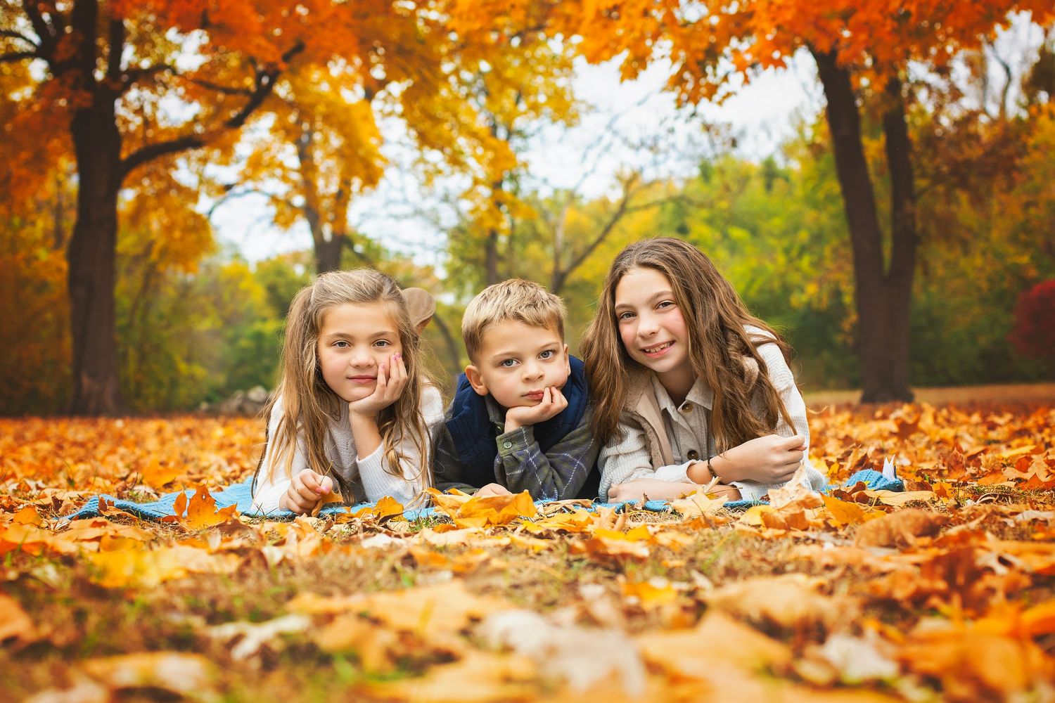 Three children lying in autumn leaves in a park with colorful fall foliage.