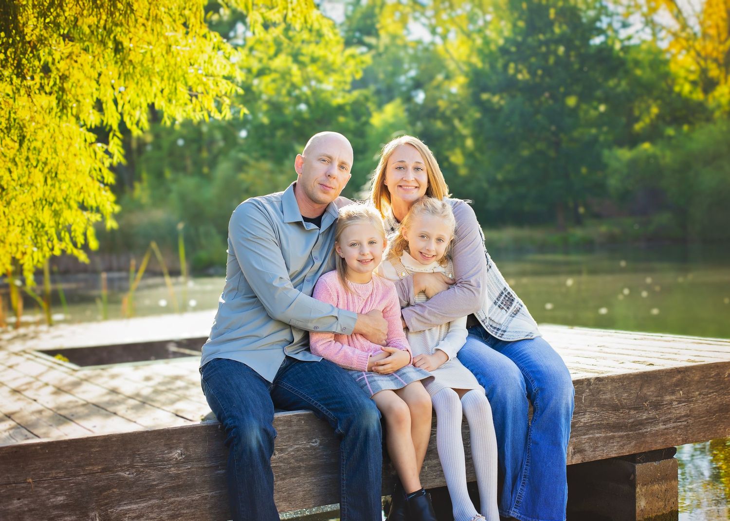 Family of four sitting on a wooden dock surrounded by golden autumn trees near a lake.