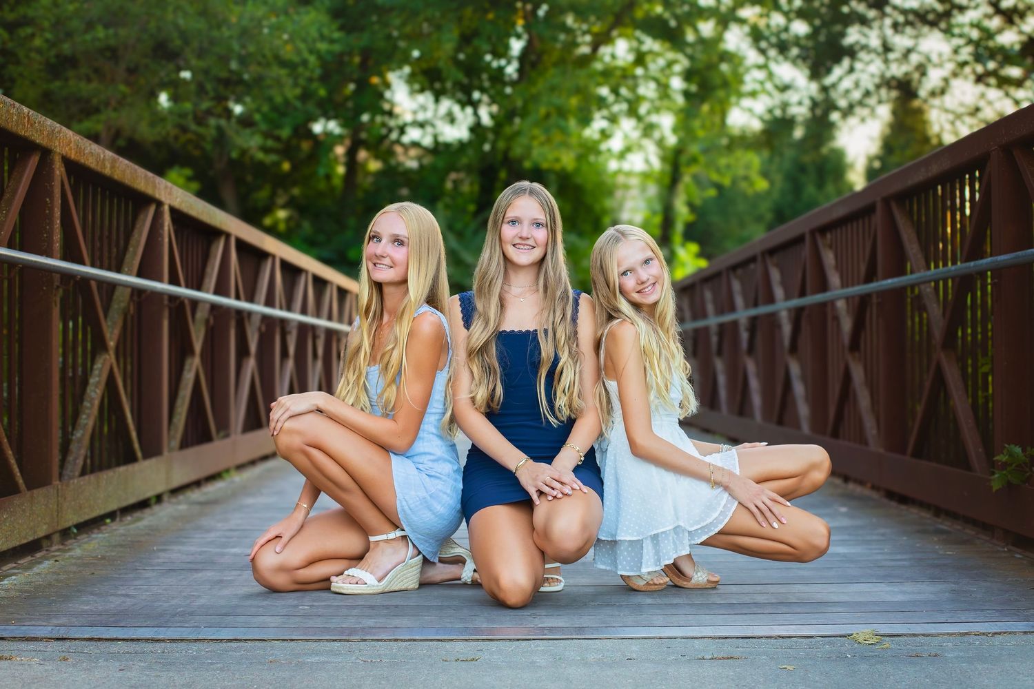 Three blonde girls pose together on a wooden bridge surrounded by lush green trees.