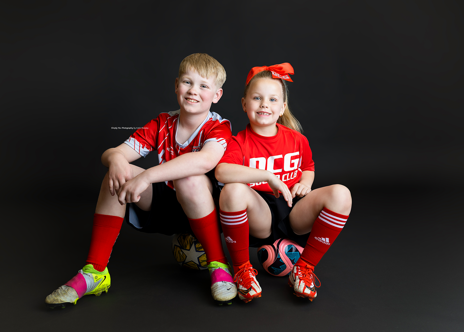 Two young children in red soccer uniforms and cleats pose together for a team photo against a dark background.
