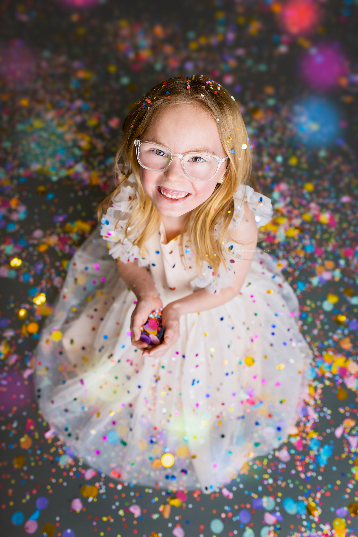 Overhead view of a smiling girl in a white polka dot dress holding confetti on a glittery studio floor.
