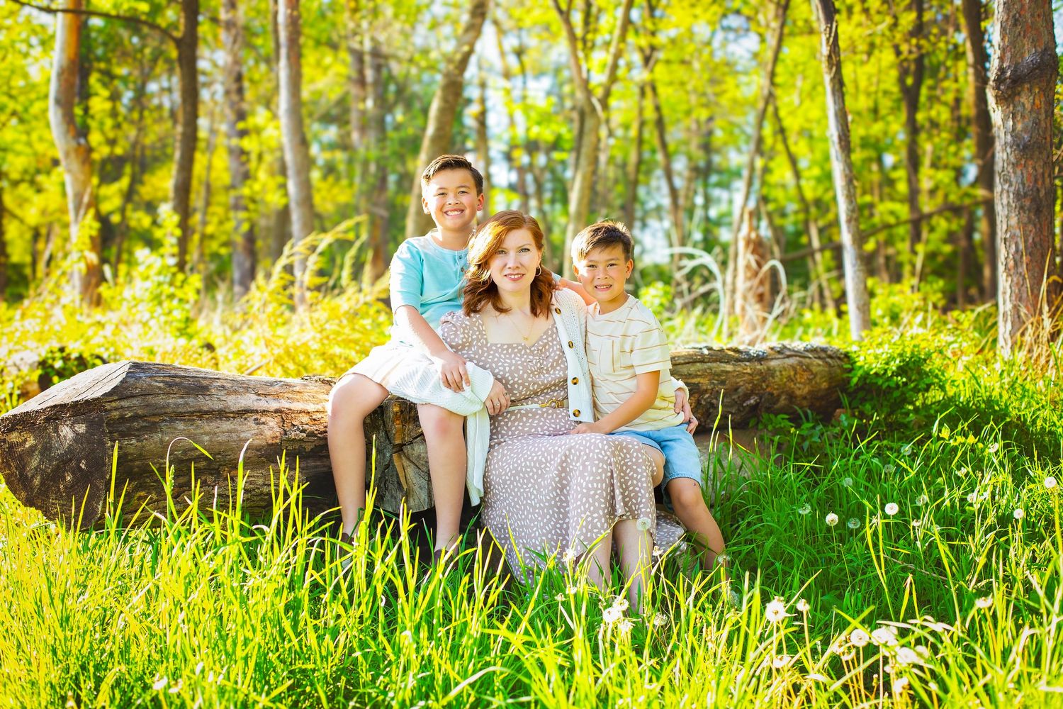 A woman and two boys sit together on a log in a sunny, lush green forest setting.